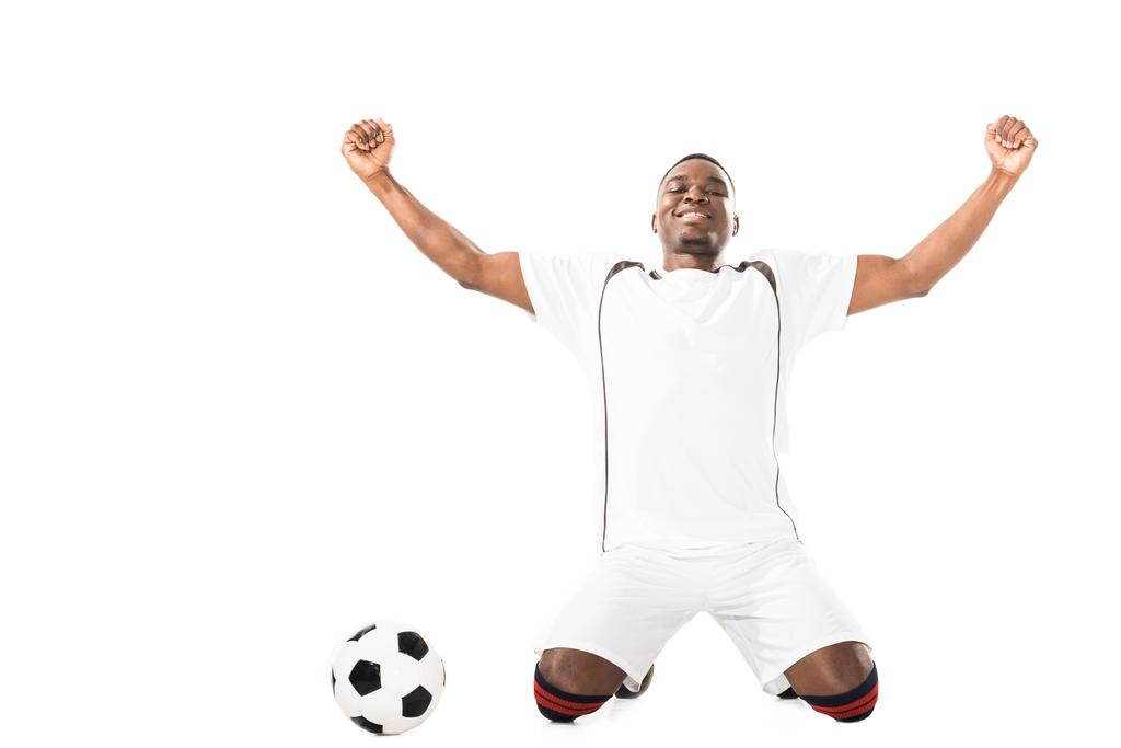 heureux jeune triomphant afro-américain joueur de football agenouillé près de ballon isolé sur blanc
   - Photo, image
