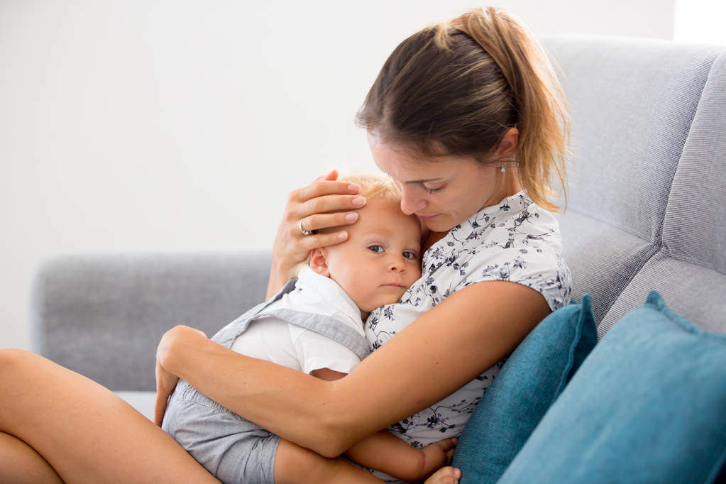 Jonge moeder, zitten thuis in zonnige woonkamer, knuffelen met haar peuter babyjongen, borstvoeding hem teder - Foto, afbeelding