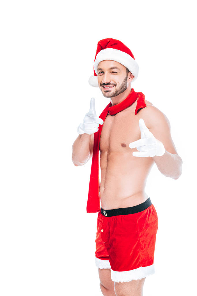 muscoloso uomo senza maglietta in cappello di Natale e sciarpa rossa strizzando l'occhio e indicando la fotocamera con le dita isolate su sfondo bianco
 - Foto, immagini