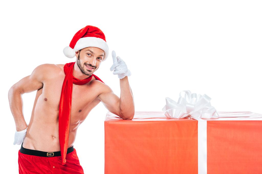 Мужчина без рубашки в рождественской шляпе и красном шарфе стоит рядом с большой подарочной коробкой и указывает на камеру, изолированную на белом фоне
 - Фото, изображение