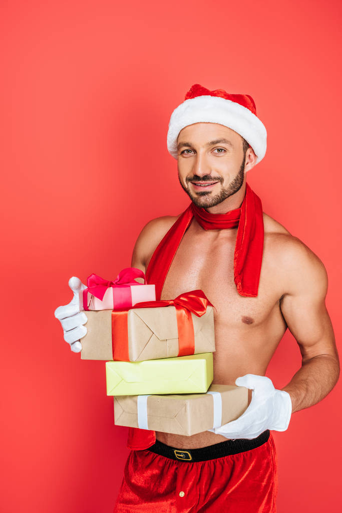 ευτυχισμένος μυώδης άνδρας shirtless σε Χριστουγεννιάτικο καπέλο και κόκκινο μαντήλι κρατώντας σωρό κουτιά δώρων που απομονώνονται σε κόκκινο φόντο  - Φωτογραφία, εικόνα
