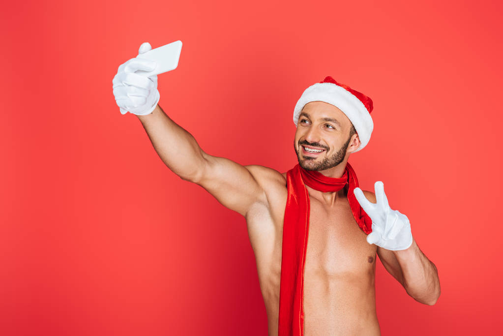χαμογελώντας μυώδης άνδρας shirtless σε Χριστουγεννιάτικο καπέλο και κόκκινο μαντήλι λήψη selfie σε smartphone και να κάνει το σημάδι ειρήνης που απομονώνονται σε κόκκινο φόντο  - Φωτογραφία, εικόνα