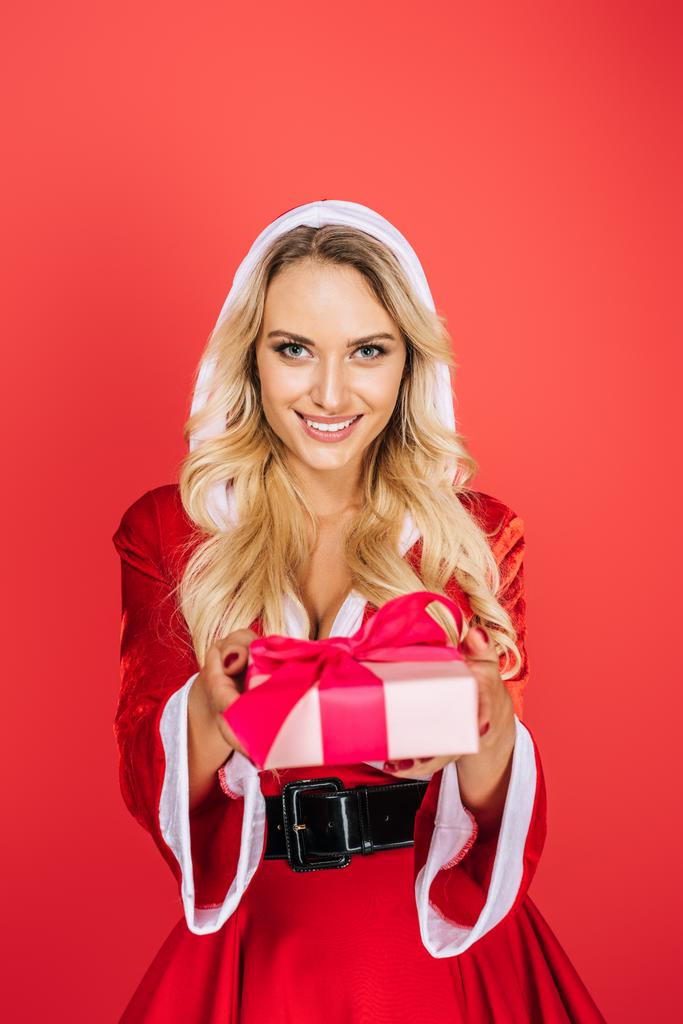 χαμογελώντας santa κορίτσι με φόρεμα Χριστούγεννα δίνοντας κουτί δώρου που απομονώνονται σε κόκκινο φόντο - Φωτογραφία, εικόνα