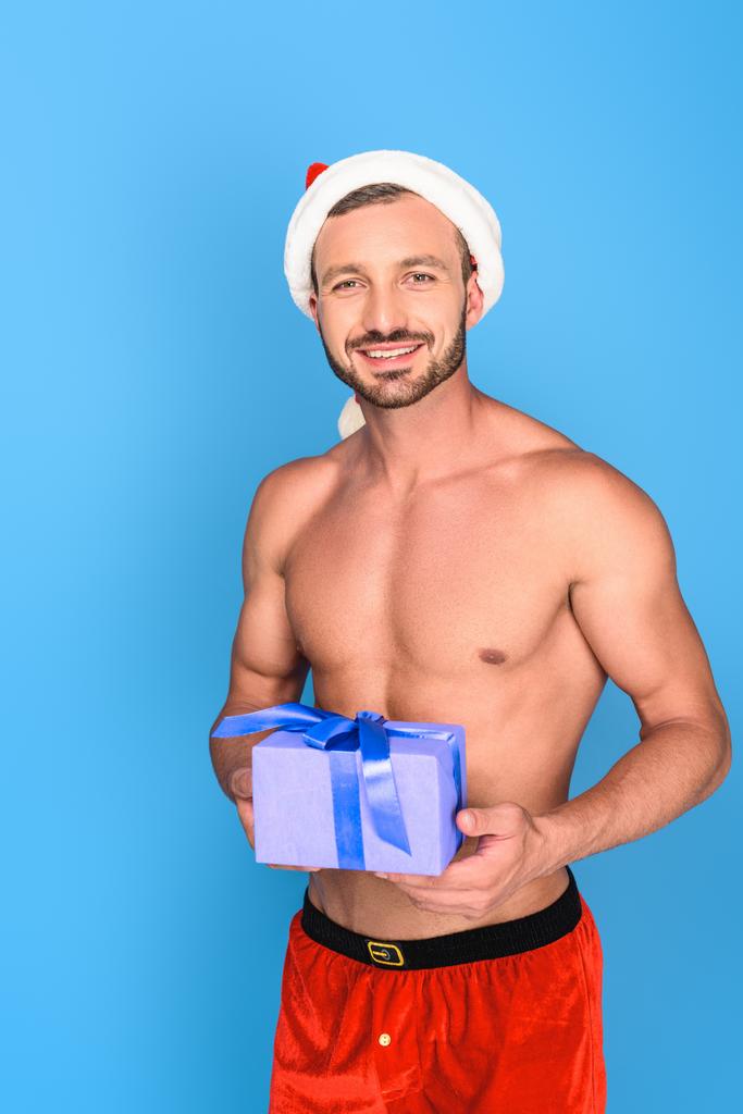 sexy sin camisa musculoso hombre en navidad sombrero celebración regalo caja aislado sobre fondo azul
 - Foto, Imagen