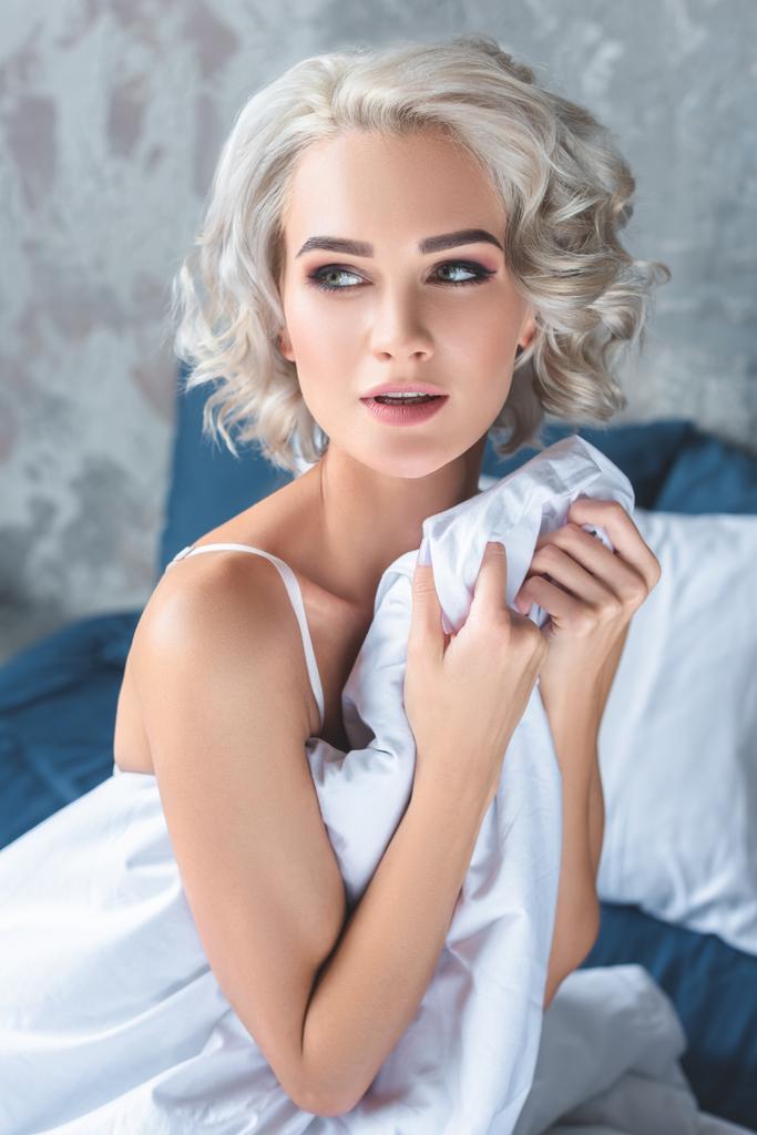 Sexy mladá žena sedící na posteli v prádle při krytí s přikrývkou a hledat dál - Fotografie, Obrázek