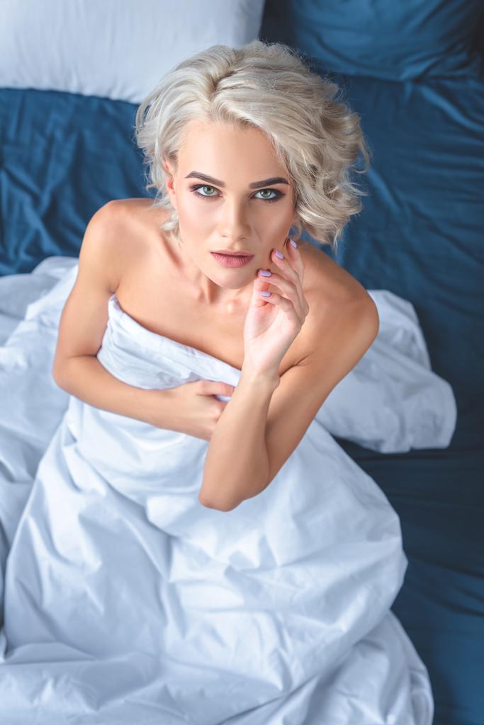 μεγάλη γωνία προβολής ελκυστική γυμνή νεαρή γυναίκα που κάθεται στο κρεβάτι, καλύπτοντας το σώμα με κουβέρτα και βλέπουν τα φωτογραφικών μηχανών - Φωτογραφία, εικόνα