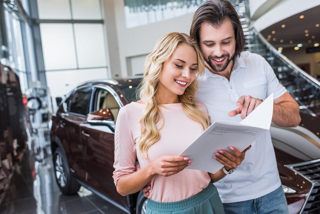 portrait of cheerful couple with catalog buying car at dealership salon - Photo, Image
