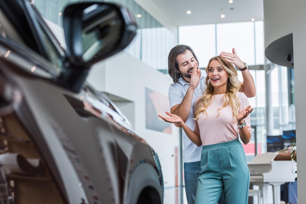 uśmiechnięty mężczyzna zaskakujące dziewczyna z nowego samochodu w salonie dealera - Zdjęcie, obraz
