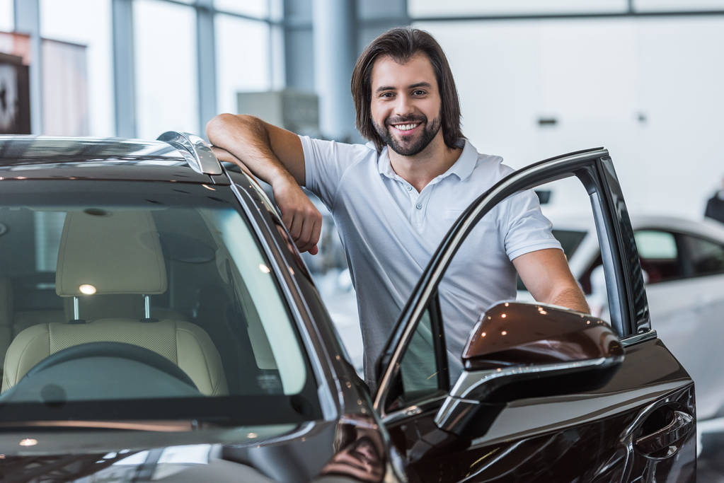 Porträt eines lächelnden Mannes, der vor einem Neuwagen im Autohaus steht - Foto, Bild