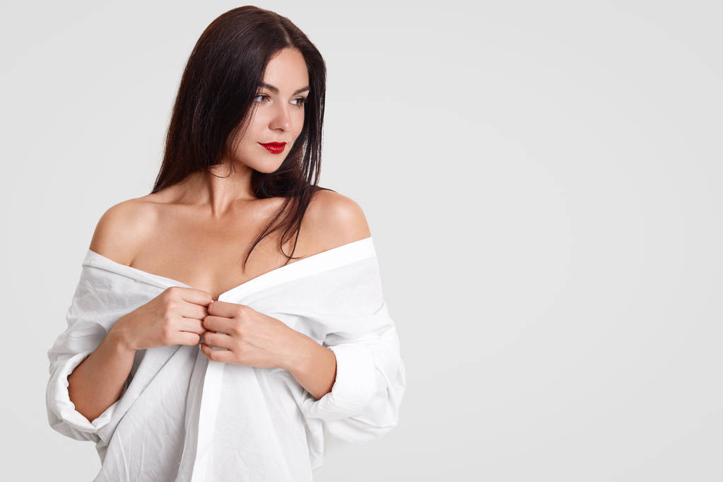 Isolated shot of pretty brunette female with sexual fit body, shows bare shoulders, wears oversized white shirt, stands against studio wall with copy space for your advertising content or text - Photo, Image