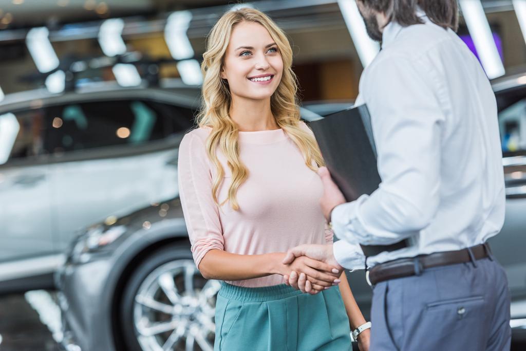 vista parcial del concesionario asistente de tienda y sonriente cliente femenino estrechando la mano en el salón de automóviles
 - Foto, Imagen