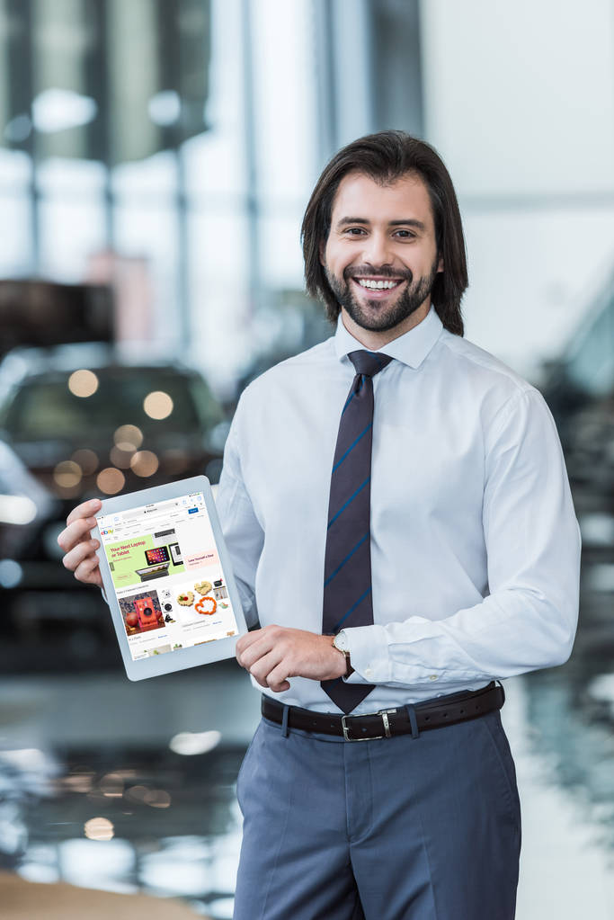 vendedor de salón de concesionarios sonriente en ropa formal que muestra la tableta con el sitio web ebay en la pantalla en las manos
  - Foto, Imagen