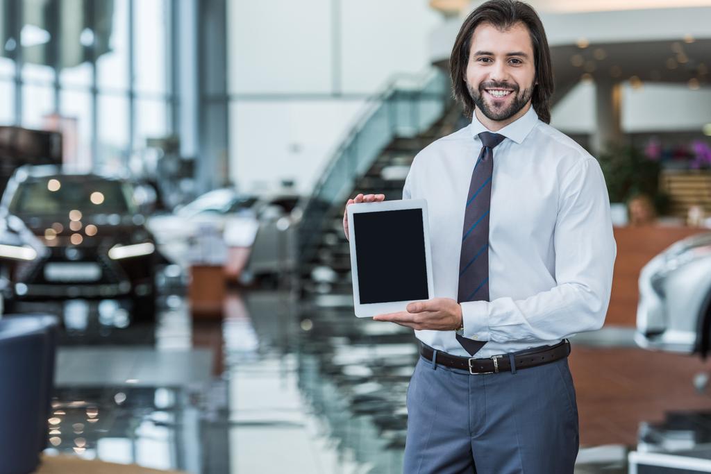 retrato del vendedor sonriente en ropa formal mostrando tableta con pantalla en blanco en el salón de concesionarios
 - Foto, Imagen