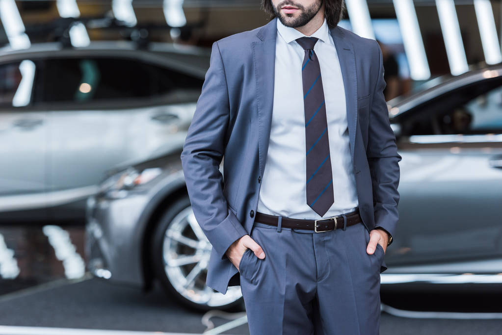 частичный взгляд бизнесмена в стильном костюме, стоящего в салоне дилера с новыми автомобилями на заднем плане
 - Фото, изображение