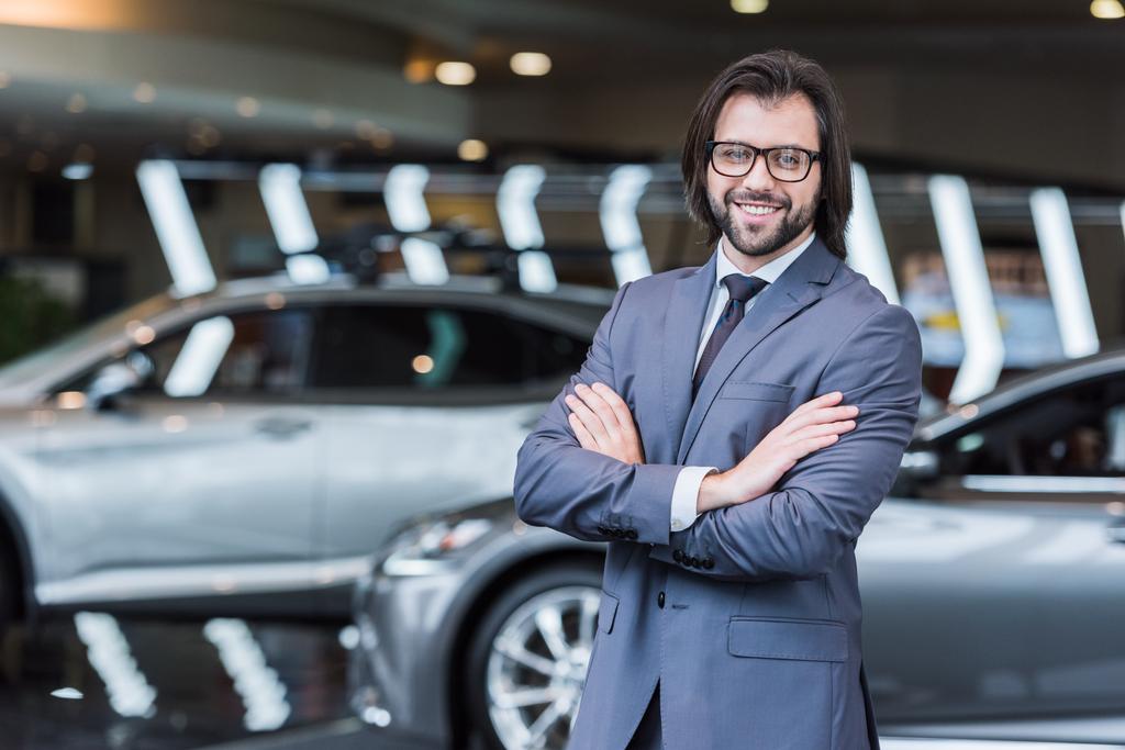 sonriente hombre de negocios con los brazos cruzados de pie en el salón de concesionarios con coches nuevos en el fondo
 - Foto, imagen