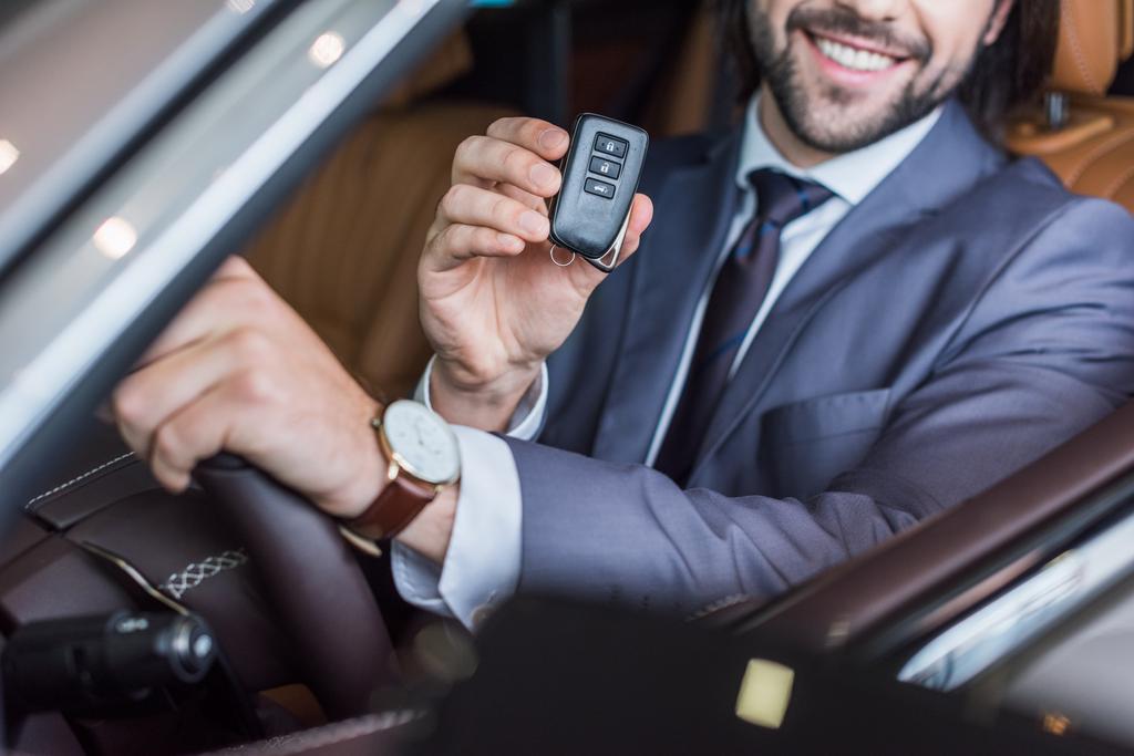 vista parcial de sonriente hombre de negocios con la llave del coche en la mano sentado en un coche nuevo en el salón de concesionarios
 - Foto, Imagen