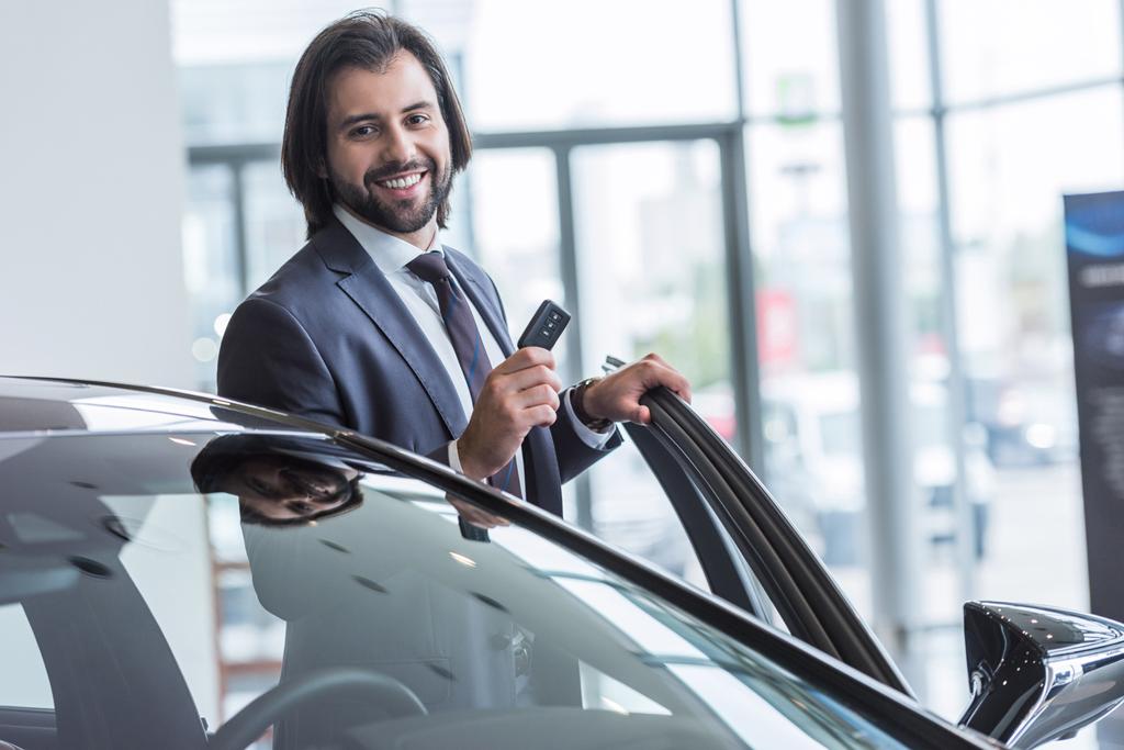 Porträt eines lächelnden Geschäftsmannes mit Autoschlüssel, der vor einem Neuwagen im Autohaus steht - Foto, Bild