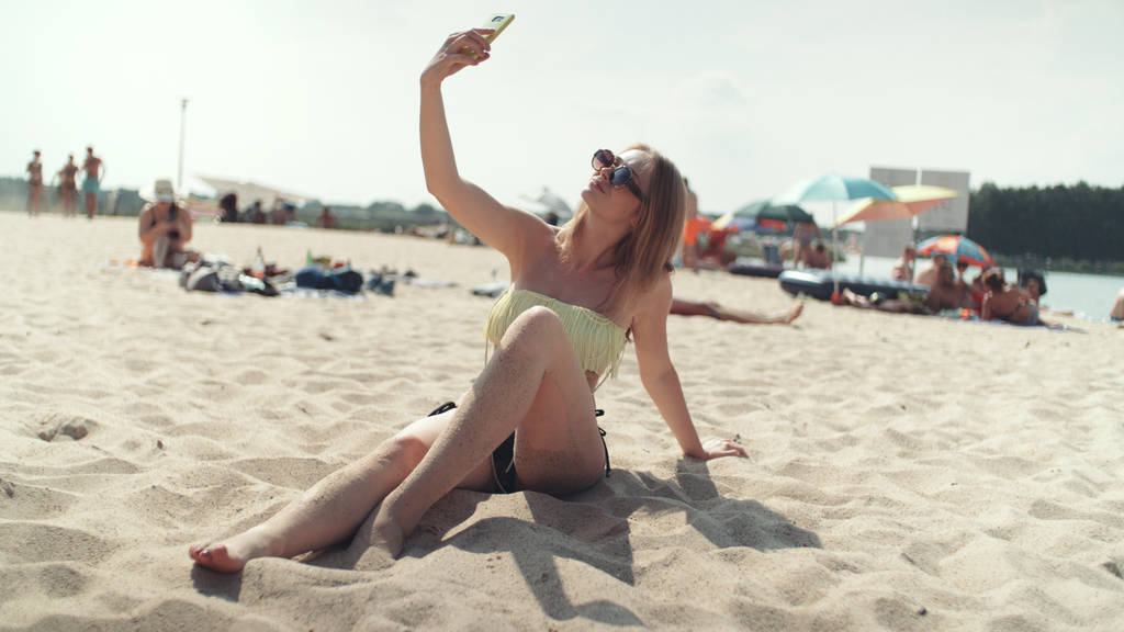 Όμορφη γυναίκα στο μπικίνι, κάθονται στην παραλία και λαμβάνοντας μια selfie. - Φωτογραφία, εικόνα