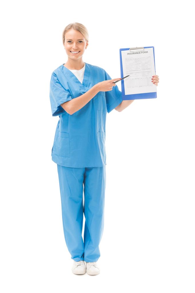 smiling young nurse pointing at clipboard with insurance form isolated on white - Photo, Image
