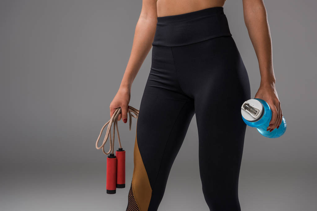 обрезанный снимок спортивной женщины в леггинсах с скакалкой и бутылкой воды на сером
 - Фото, изображение