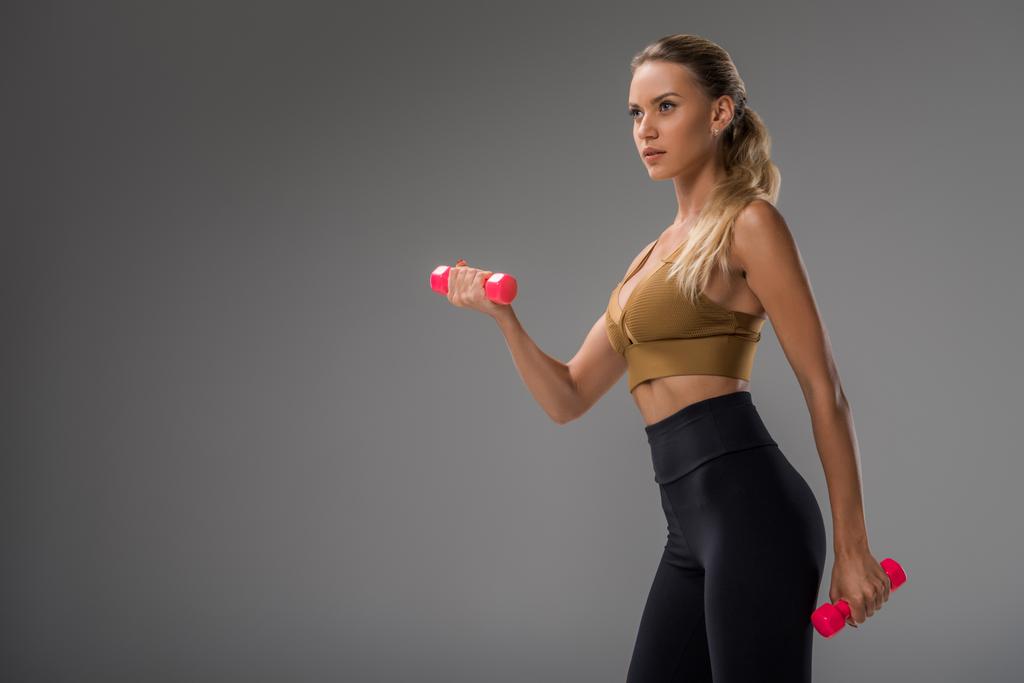 атлетичная молодая женщина, тренирующаяся с гантелями на сером
 - Фото, изображение