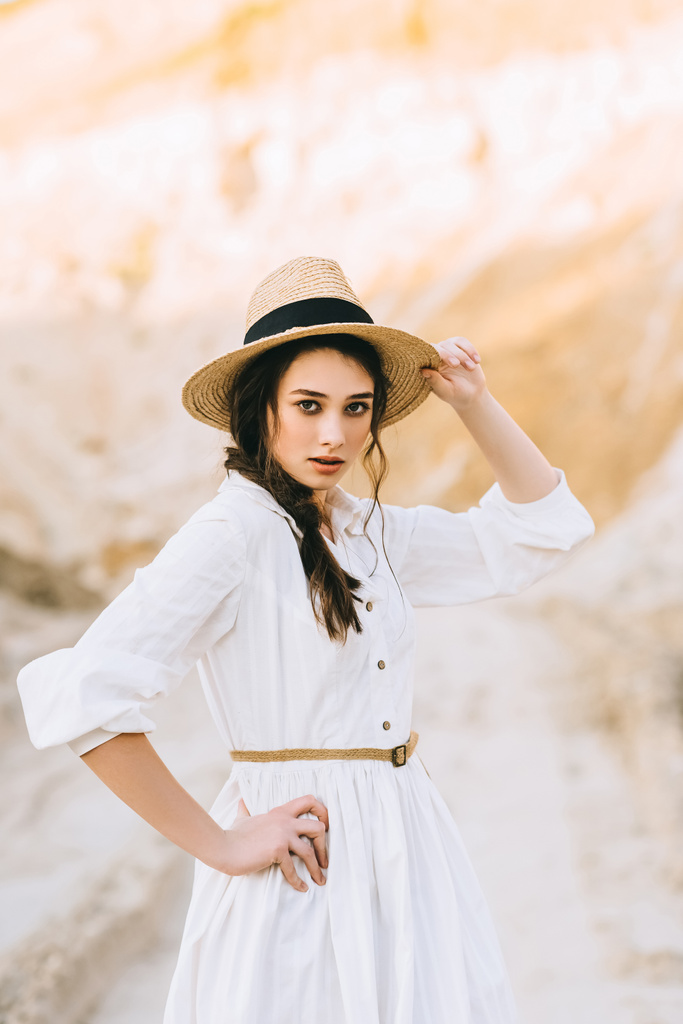 ελκυστική κοπέλα ποζάρει σε κομψό φόρεμα και ψάθινο καπέλο σε αμμώδη φαράγγι - Φωτογραφία, εικόνα