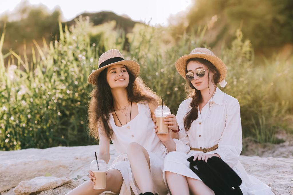 beautiful smiling friends in straw hats with coffee latte sitting on ground - Photo, Image