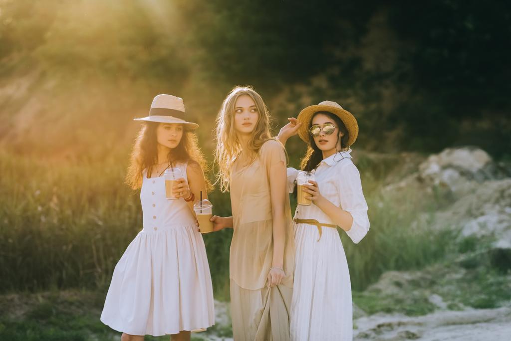 елегантні молоді жінки в солом'яних капелюхах тримають чашки з кавовим латте і позують з сонячним світлом
 - Фото, зображення