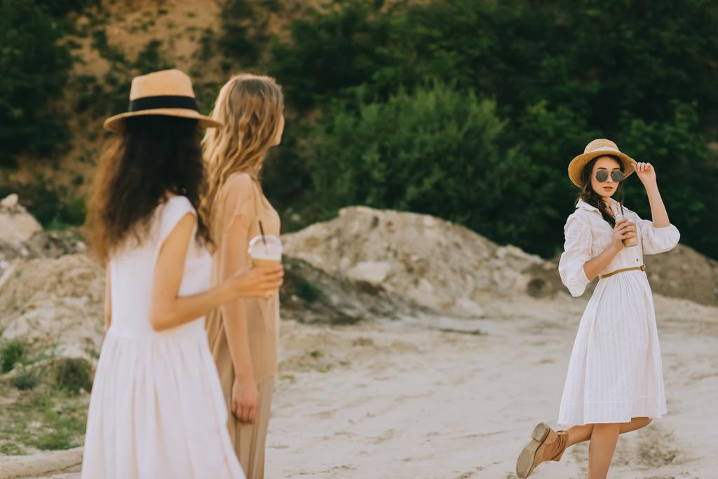 meninas elegantes atraentes em chapéus de palha andando na natureza com xícaras de plástico de café latte
 - Foto, Imagem