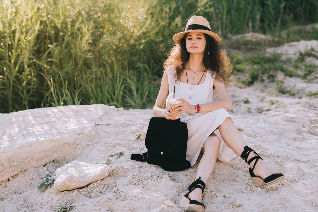 привлекательная кудрявая девушка с кофе латте и рюкзаком сидя на земле
 - Фото, изображение