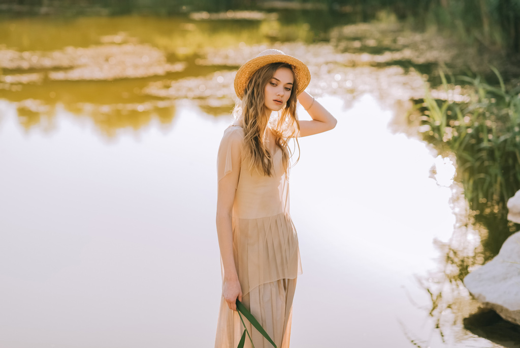 mooi blonde meisje in elegante jurk en stro hoed poseren in de buurt van lake - Foto, afbeelding