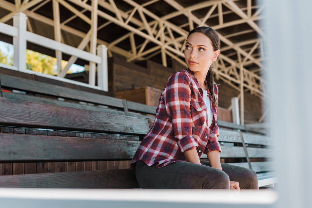 beautiful cowgirl in checkered shirt sitting on bench at ranch stadium and looking away - Photo, Image