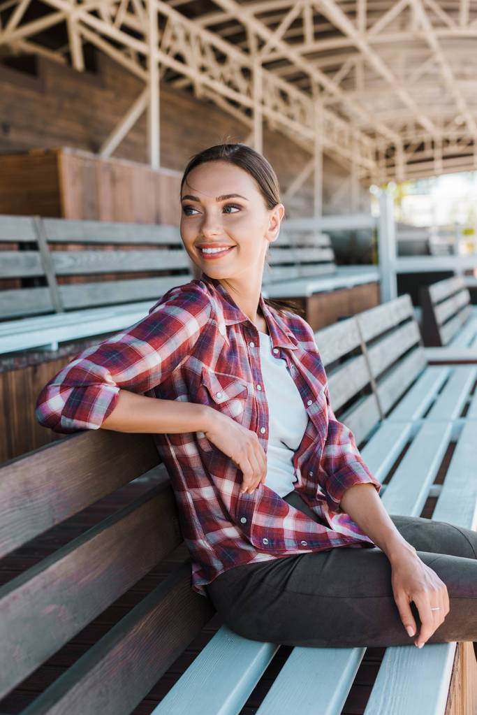donna attraente sorridente in camicia a scacchi seduta sulla panchina allo stadio ranch e guardando altrove
 - Foto, immagini