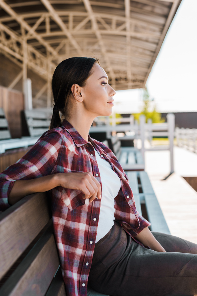 Seitenansicht des schönen Cowgirls im karierten Hemd, das auf der Bank im Ranch-Stadion sitzt und wegschaut - Foto, Bild