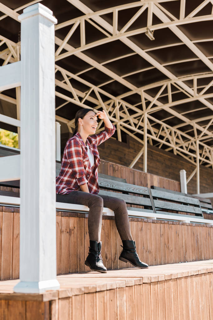attractive cowgirl in checkered shirt sitting on bench at ranch stadium and looking at something - Photo, Image