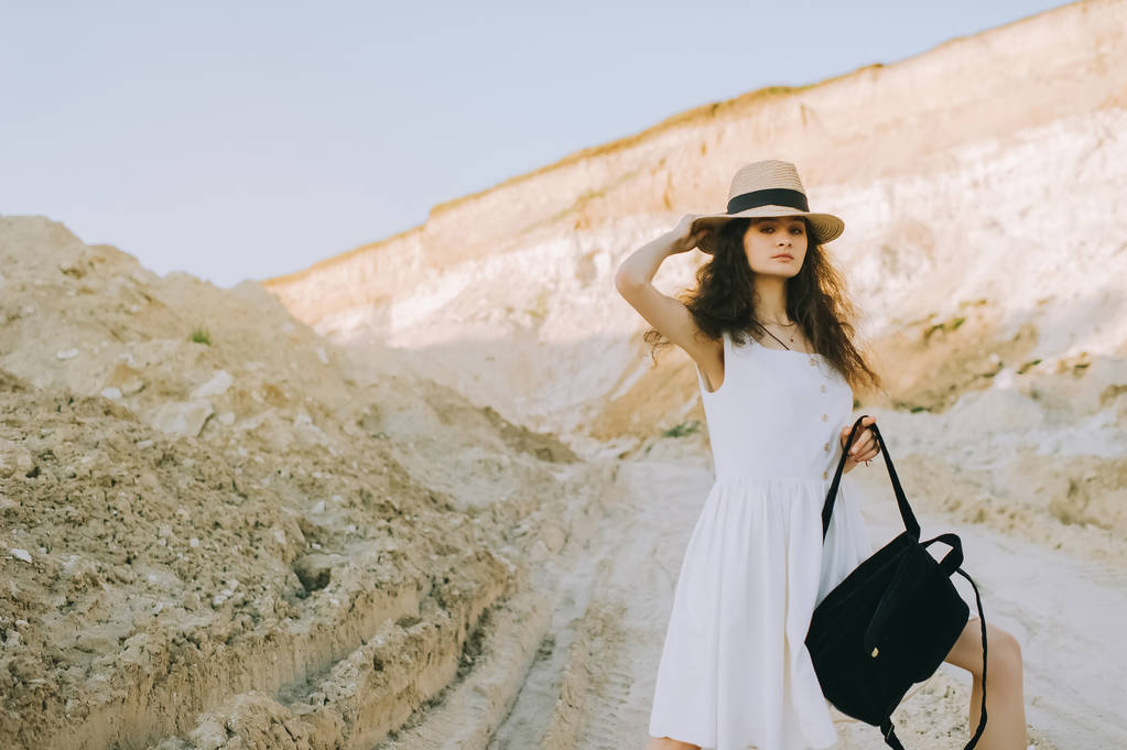 beautiful curly girl in white dress and straw hat posing with backpack in sandy canyon - Photo, Image