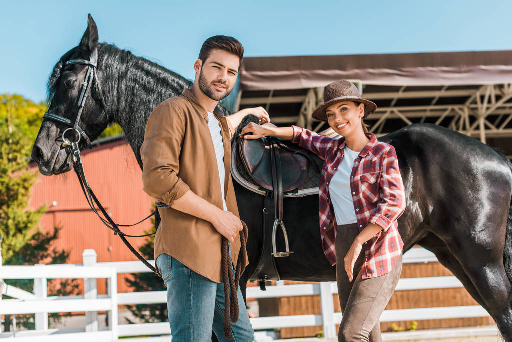 vista angolo basso di cowboy e cowgirl in piedi vicino al cavallo al ranch e guardando la fotocamera
  - Foto, immagini
