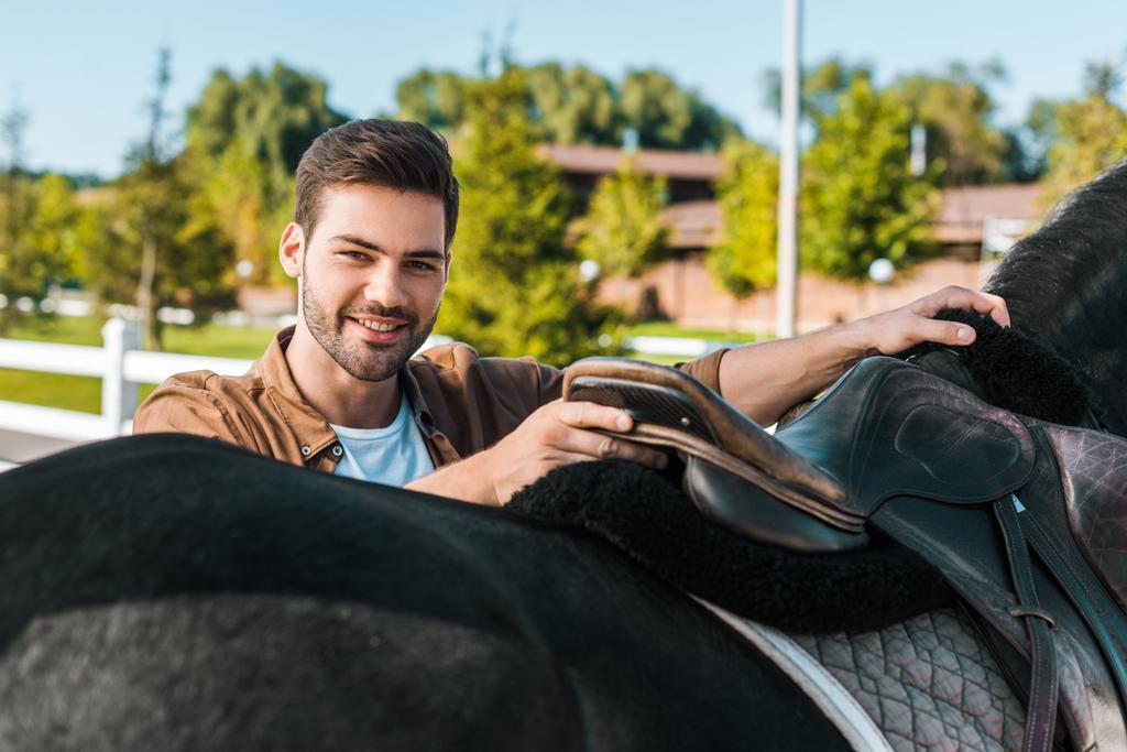 усміхнений красивий чоловічий кінний фіксуючий сідло коня на ранчо
 - Фото, зображення