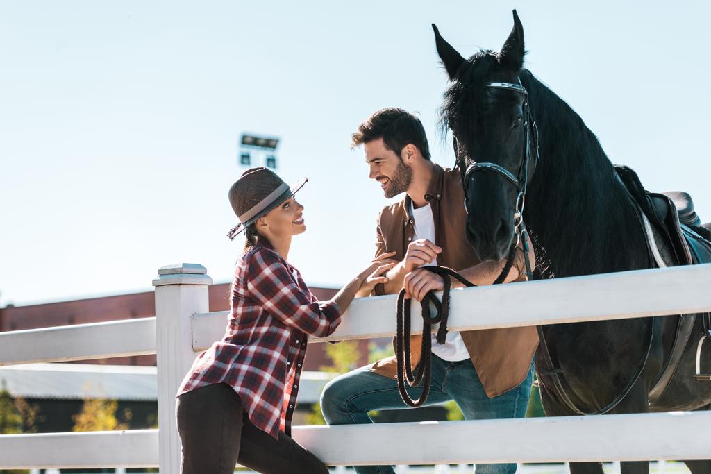 улыбающиеся наездники женского и мужского пола, стоящие у забора с лошадьми и смотрящие друг на друга на ранчо
 - Фото, изображение
