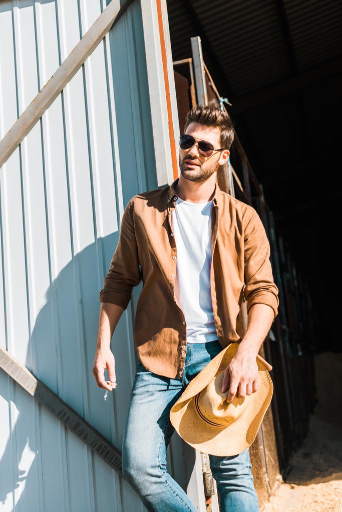 hombre guapo en gafas de sol sosteniendo cigarrillo y sombrero, apoyado en la pared en el rancho
 - Foto, imagen