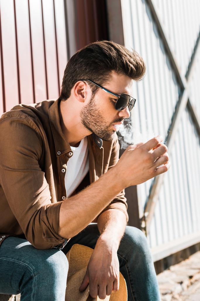 side view of handsome cowboy in sunglasses sitting and smoking cigarette at ranch - Photo, Image