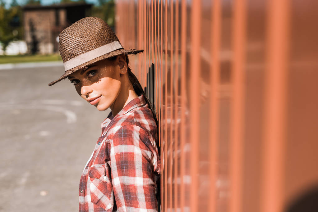 attractive cowgirl in checkered shirt and hat leaning on brown wall at ranch and looking at camera - Photo, Image