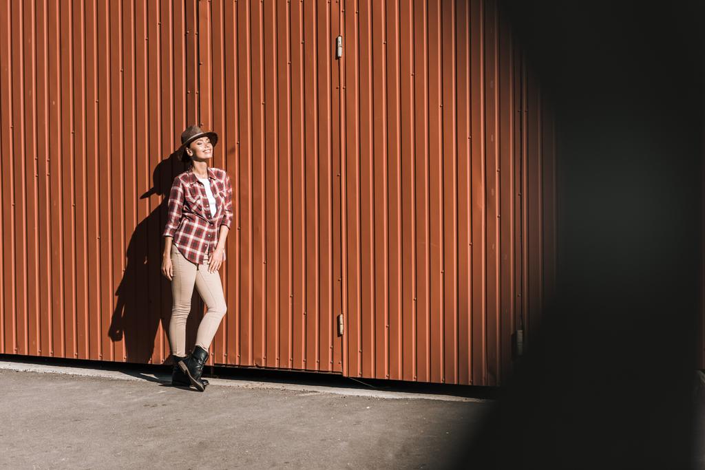 lachende aantrekkelijke vrouw in geruite shirt en muts leunend op bruin muur op ranch - Foto, afbeelding