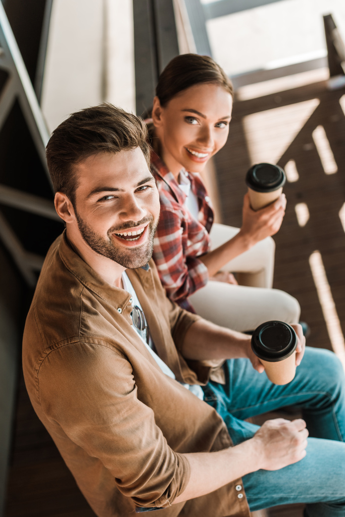 Hochwinkelaufnahme eines lächelnden Mannes und einer lächelnden Frau, die mit Kaffee to go auf der Ranch sitzen und in die Kamera schauen - Foto, Bild