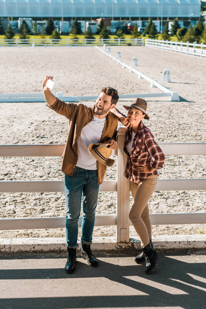 glimlachend stijlvolle cowboy en cowgirl in casual kleding nemen selfie met smartphone op ranch - Foto, afbeelding