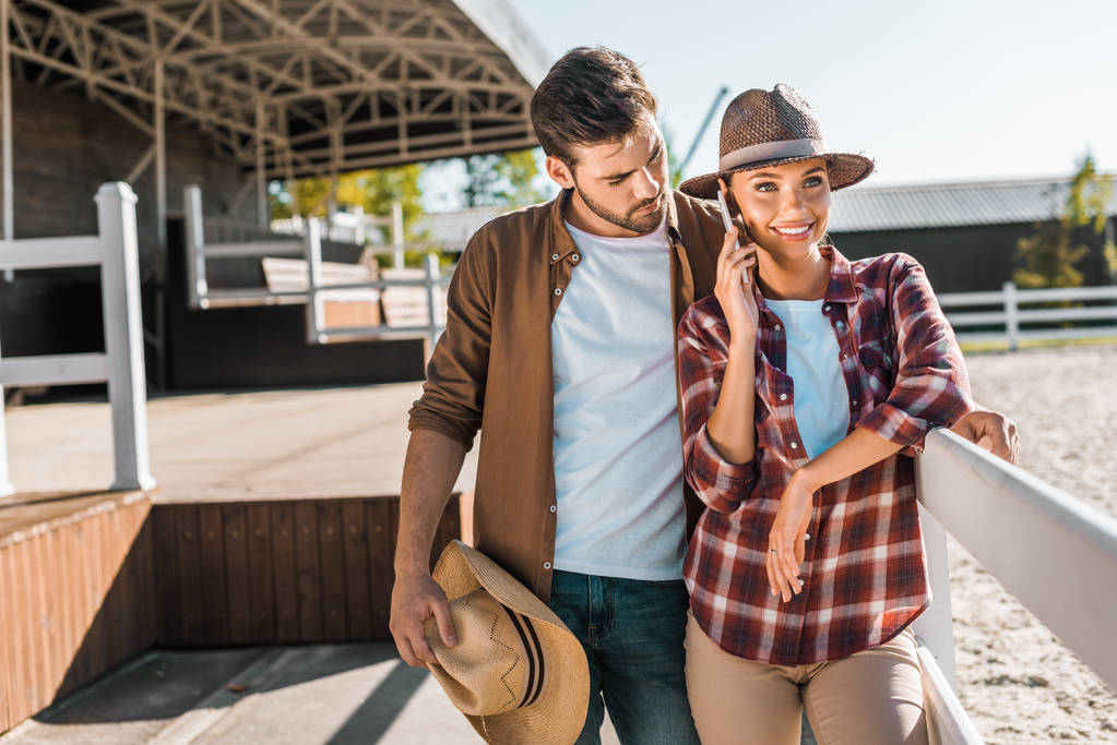 tyylikäs cowboy ja cowgirl rento vaatteita seisoo aidan lähellä tilalla, nainen puhuu älypuhelimella
 - Valokuva, kuva