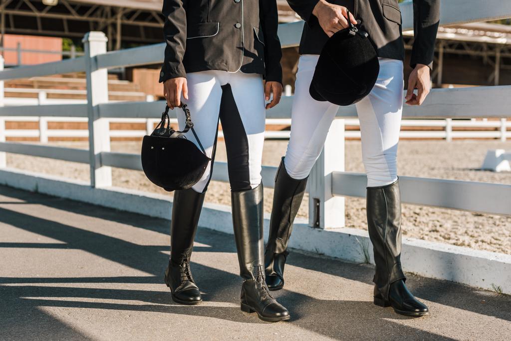 bijgesneden afbeelding van ruiters in professionele kleding lopen in de buurt van hek en houdt van paardrijden helmen op ranch - Foto, afbeelding