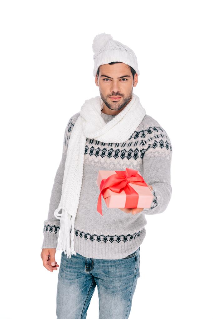 schöner junger Mann in Pullover, Schal und Mütze mit Geschenkbox und Blick in die Kamera isoliert auf weiß   - Foto, Bild