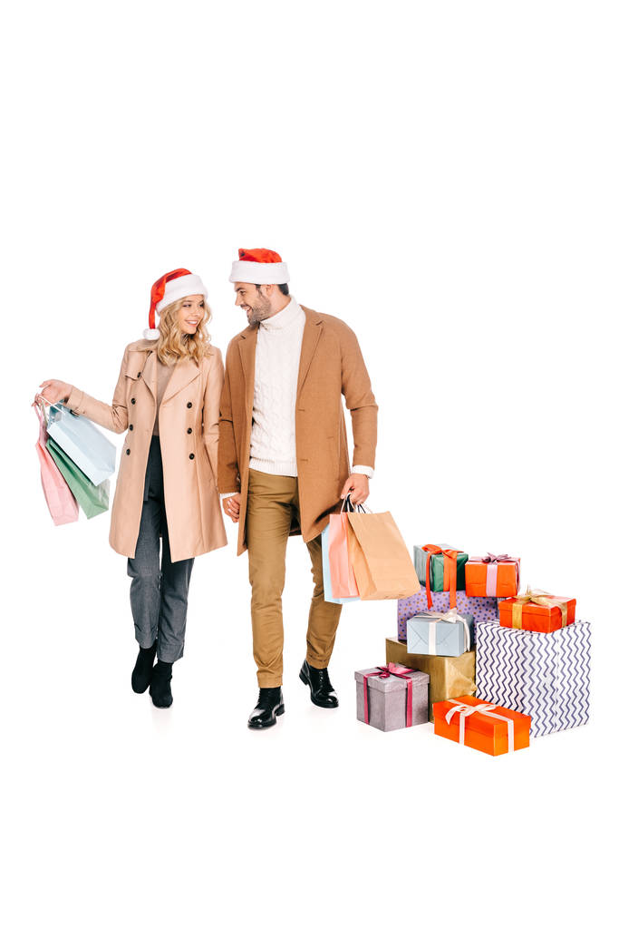 улыбающаяся молодая пара в шляпах Санты, держащая сумки для покупок и подходя к подарочным коробкам, изолированным на белом
 - Фото, изображение