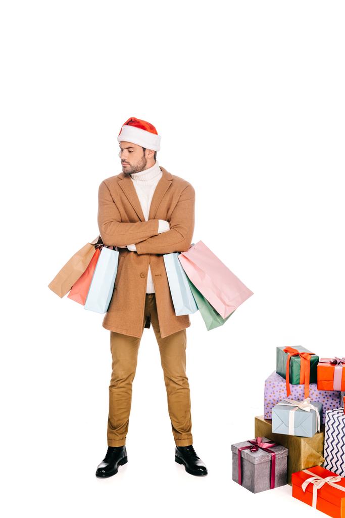 young man in santa hat holding shopping bags while standing near christmas presents isolated on white  - Photo, Image